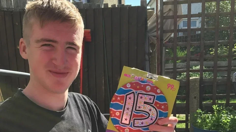 A boy with blonde hair and a black top. He holds a birthday badge.